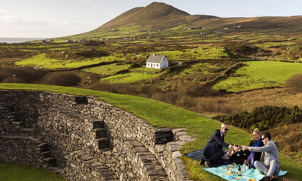 Reconnect in Ireland - Image shown here shows two men and a woman having a champagne picnic on an exterior wall at Cahergal Stone Fort, County Kerry.