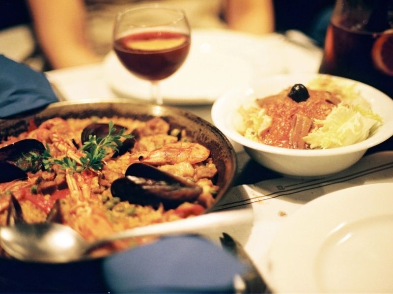 Spain's food and wine offerings range from paellas to rich stews. Image here shows pan of paella with glass of wine in background. Photo is by jroberblack | Canva.