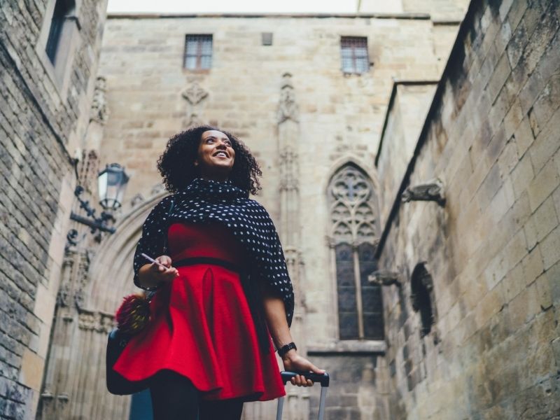 Spain is safe to travel to. Image shows a woman in a red dress smiling and pulling a suitcase through Barcelona's Gothic Quarter. Photo is by martindm | Canva.